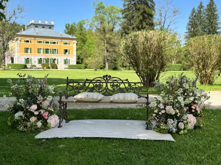 Un banc en fer forgé, décoré de coussins et de fleurs pour un mariage mis en place dans le splendide jardin verdoyant de la Villa Palladienne visible en arrière plan